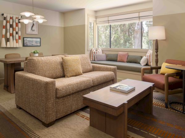 Cozy living room with a beige sofa, wooden coffee table, windowed nook, mid-century chair, dining area, and warm, neutral decor end.