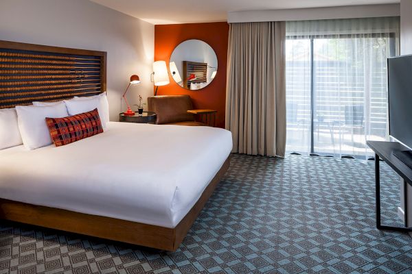 A modern hotel room with a large bed, desk with chair, round mirror, wall-mounted TV, and sliding glass doors leading to a balcony.