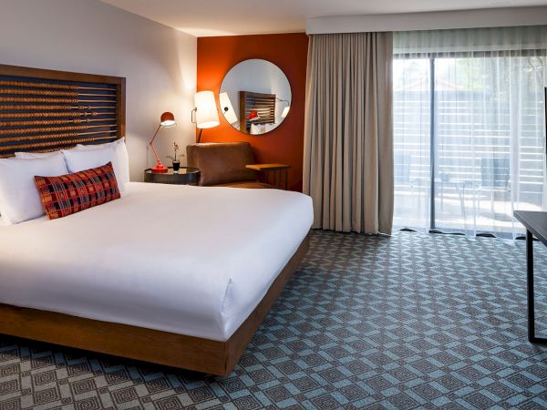 A modern hotel room with a large bed, desk with chair, round mirror, wall-mounted TV, and sliding glass doors leading to a balcony.