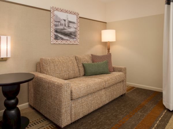Cozy hotel-style living area with a beige sofa, green throw pillow, wall lamp, framed artwork, round side table, and a curtain in a softly lit beige room.