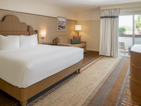 A hotel room with a large bed, two bedside lamps, a seating area, desk and balcony doorway; neutral tones and patterned carpet.