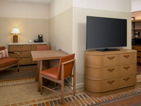 A modest hotel suite with a wooden dresser, a flat-screen TV, a rectangular table and chairs, a sofa, a lamp, and a small coffee station in a beige-toned room.