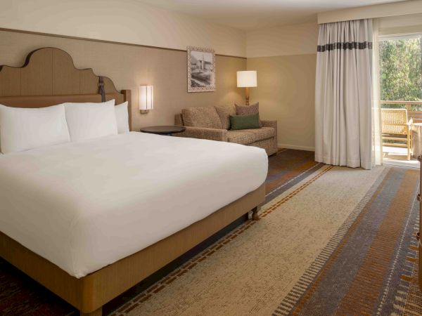 A hotel room with a king bed, sofa, nightstand lamps, and a sliding door to a balcony, all in neutral tones.