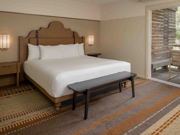 A hotel room with a king bed, two bedside tables, wall lamps, a bench at the foot, and a sliding glass door opening to a balcony.