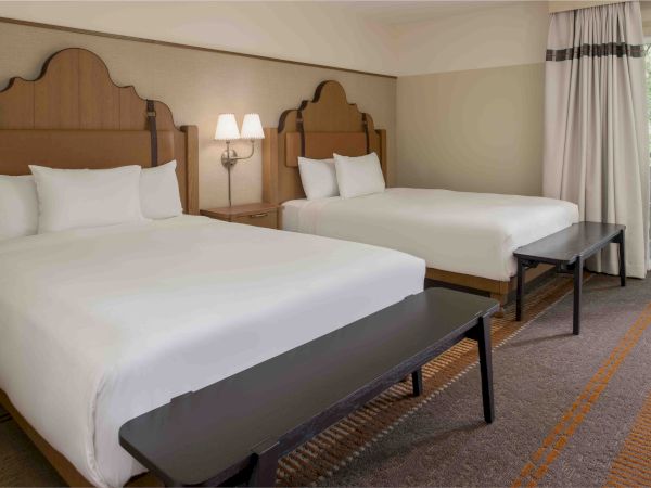 Two double beds with white linens in a hotel room, a bench at each foot, bedside lamps, and a window with curtains by the door.