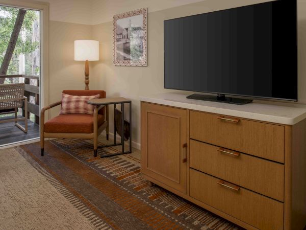 A hotel room with a dresser and a mounted TV, a chair with a side table, a floor lamp, and a doorway to a balcony or outdoor seating area.