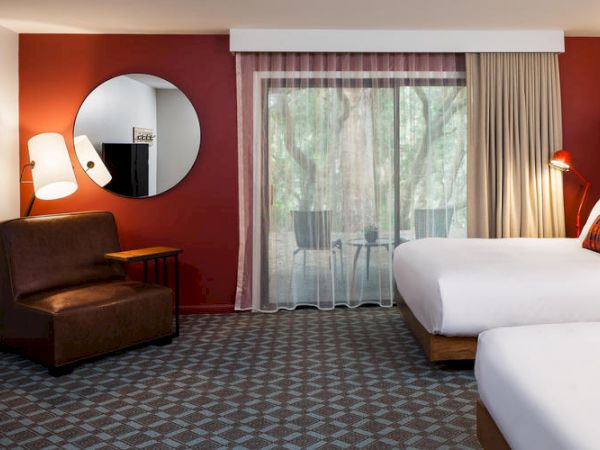 A hotel room with two beds, a sofa, a round mirror, a floor lamp, and a sliding glass door leading to a balcony with outdoor seating.