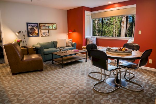 A cozy living room with a red accent wall, a green sofa, a round dining table with chairs, a large window, and wall art.