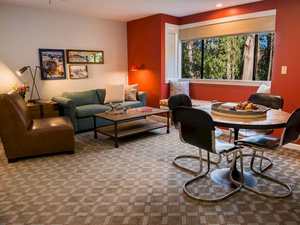 A cozy living room with a red accent wall, a green sofa, a round dining table with chairs, a large window, and wall art.