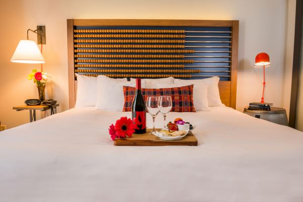 A cozy bedroom setup with a plush bed adorned with flowers, wine, glasses, and snacks on a tray, flanked by stylish lamps on both sides.