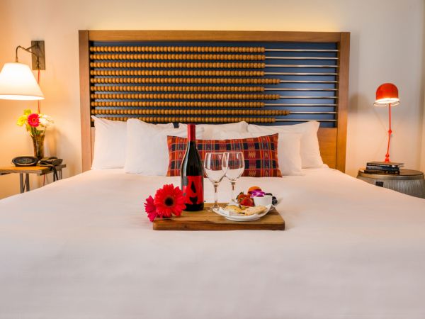 A cozy bedroom setup with a plush bed adorned with flowers, wine, glasses, and snacks on a tray, flanked by stylish lamps on both sides.