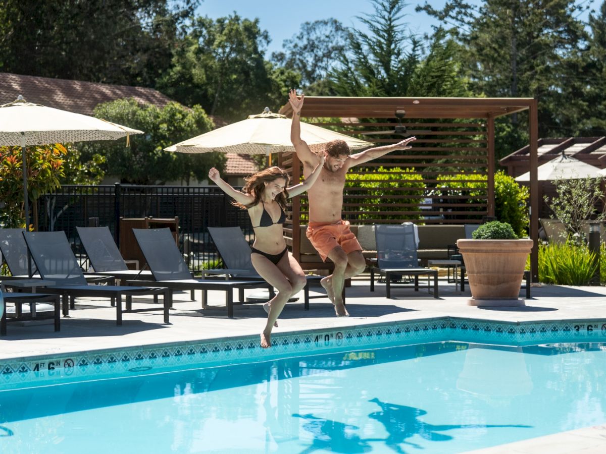Two people are jumping into a pool from the edge, a sunny poolside with lounge chairs and umbrellas in the background.