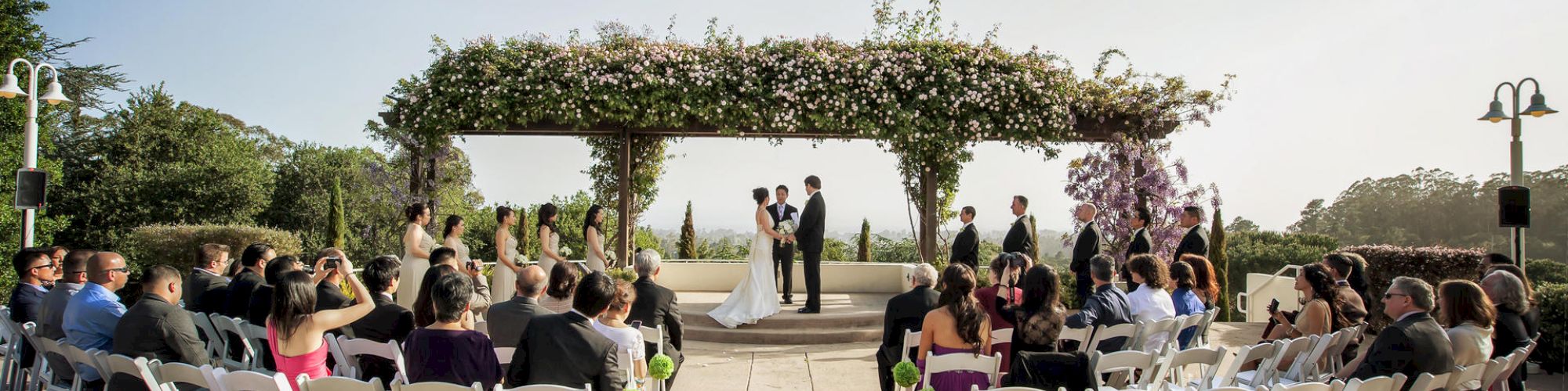 A wedding ceremony takes place outdoors, with the bride and groom standing under an arbor adorned with flowers, surrounded by seated guests.