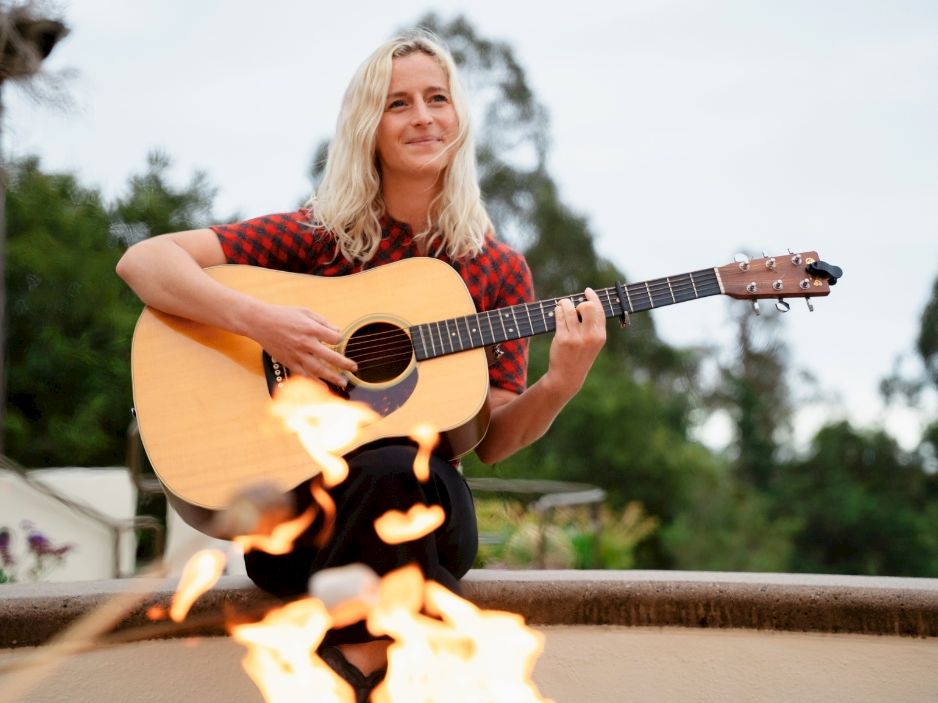 A woman sits with an acoustic guitar by a fire, outdoors, smiling, wearing a red plaid shirt; a relaxed, scenic scene with flames.