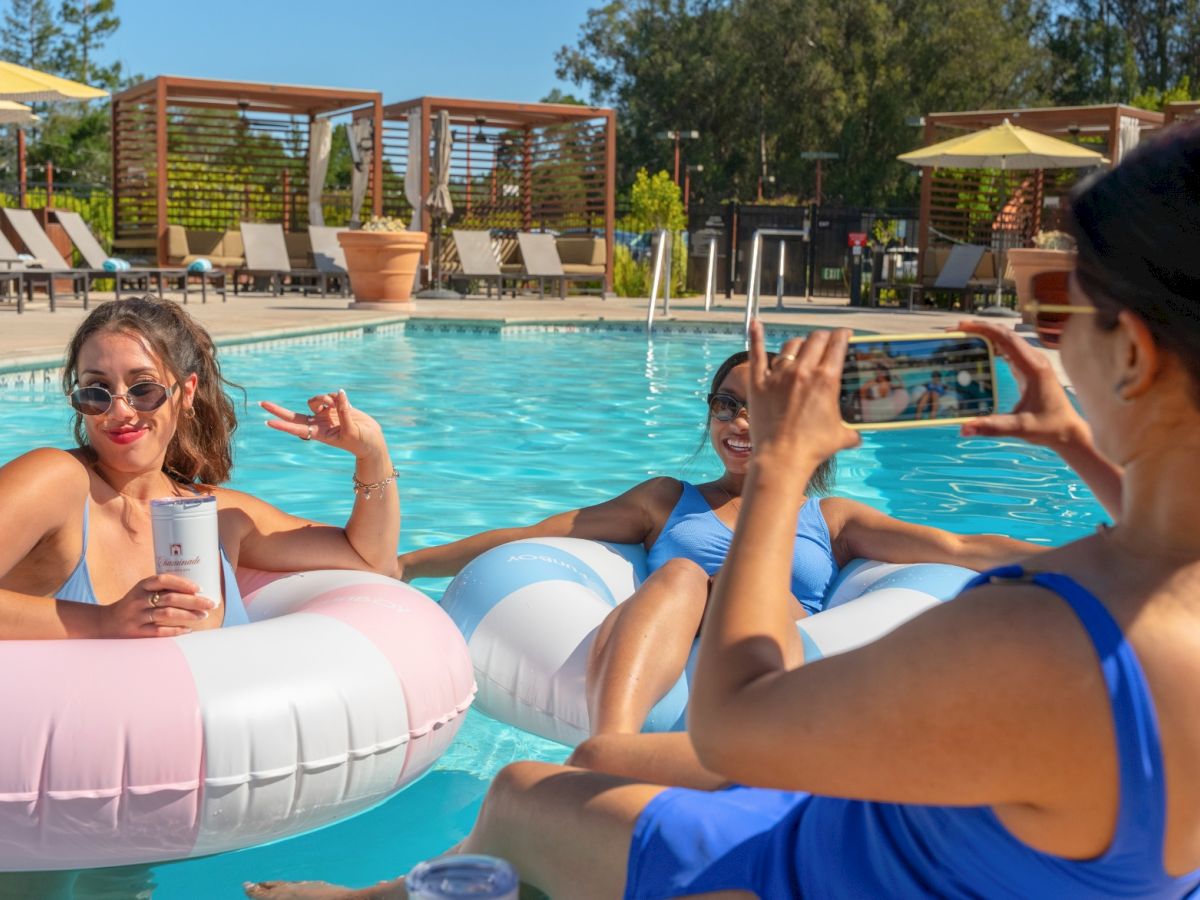 Three women in swimsuits lounge on pool floats, one taking a photo with her phone as others relax by a sunny poolside.