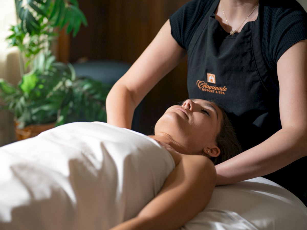 A person lies face up on a massage table, covered with a white sheet, while a practitioner in a black shirt performs a neck or head massage.