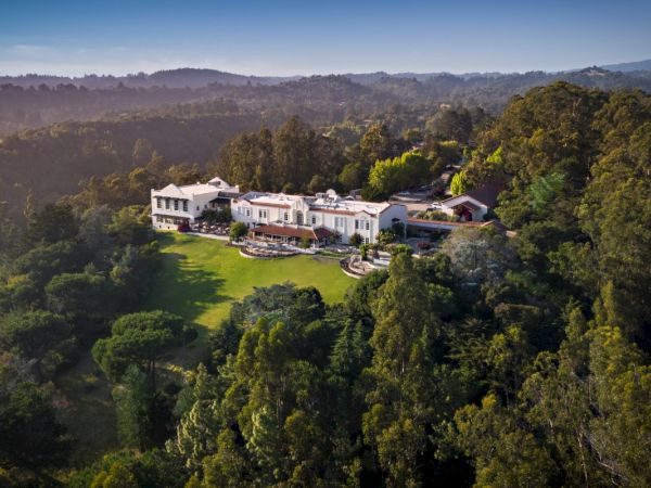 A large, luxurious mansion is nestled in a lush, green landscape with rolling hills and dense trees surrounding it.