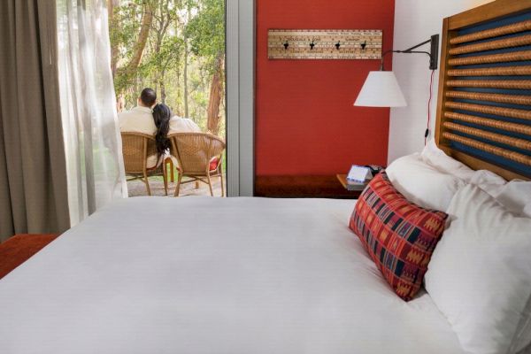 The image shows a cozy hotel room with a neatly made bed, a colorful pillow, and a person sitting outside on chairs, looking out into a forested area.