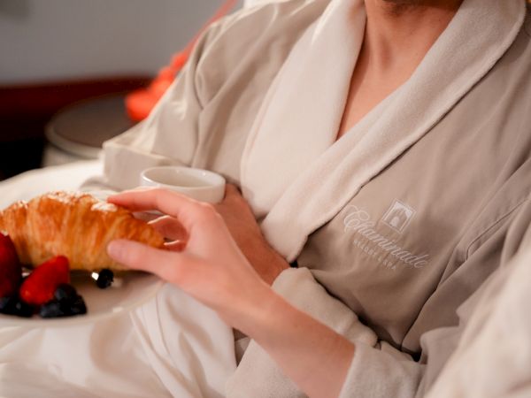 People in robes enjoying breakfast in bed, featuring a croissant, berries, and a cup of coffee on a white plate.