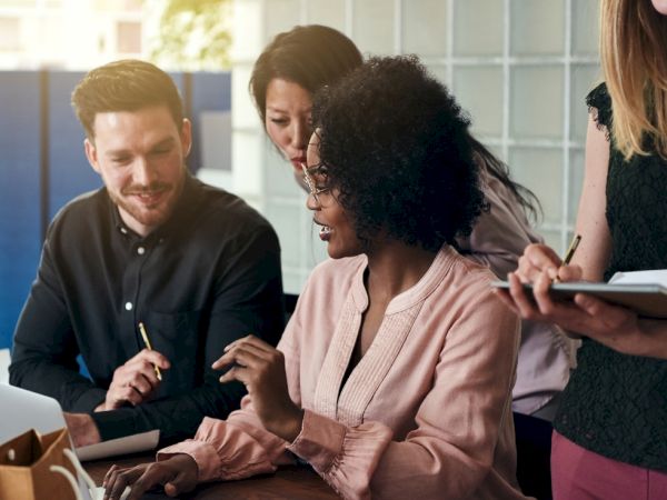 A diverse group collaborates at a table, one woman points at a laptop as others listen, discuss, and share ideas in a bright office.