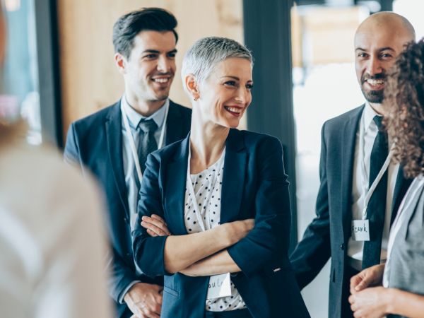 A group of people in business attire are smiling and engaging in conversation indoors, suggesting a professional or networking event.