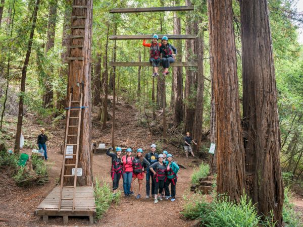 A group of people wearing helmets and harnesses are gathered in a forest, some on a rope course and others posing for a photo on the ground.