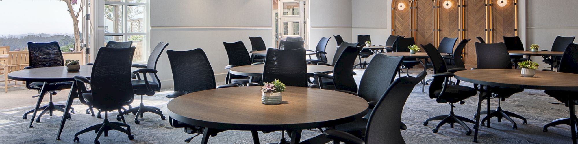 The image shows a modern conference room with round tables, black chairs, large circular ceiling lights, and wooden ceiling beams.