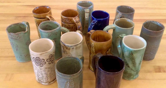 The image shows an assortment of 15 handmade ceramic mugs in various colors and designs, arranged on a wooden countertop.