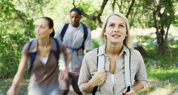 Three people are hiking through a forested area, with the woman in the front looking upward and two people behind her.