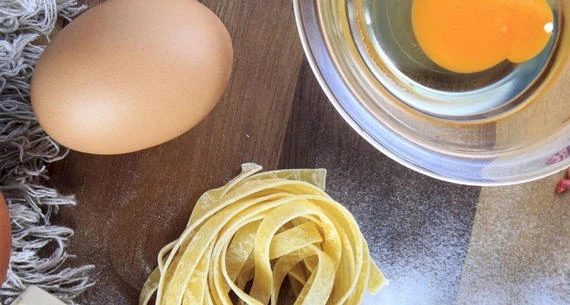 The image shows an egg, a bowl with a cracked egg, and some uncooked pasta noodles on a wooden surface.