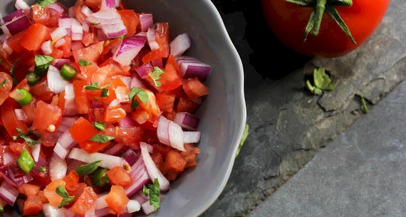 A bowl of fresh salsa made from diced tomatoes, red onions, jalapenos, and cilantro is next to a whole tomato on a dark surface.