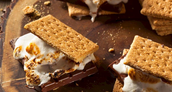 The image shows several s'mores with graham crackers, roasted marshmallows, and melted chocolate on a wooden surface. Some crumbs are visible.