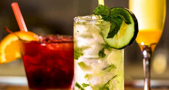 The image shows three different cocktails: a red drink with an orange slice, a clear drink with mint and cucumber, and a yellow drink in a flute glass.