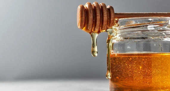 This image shows a honey dipper with honey dripping from it, placed on top of an open honey jar against a plain background.