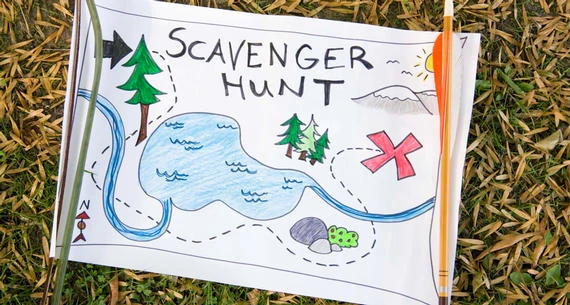 A colorful hand-drawn scavenger hunt map lies on grass, featuring a lake, trees, a red “X,” and an arrow compass pointing north.