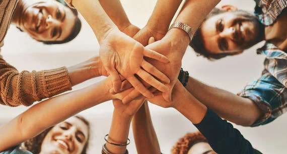 A group of people are standing in a circle with their hands joined together in the center, suggesting teamwork or unity.