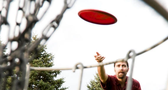 A person is throwing a red disc towards a disc golf basket outdoors, with trees visible in the background.