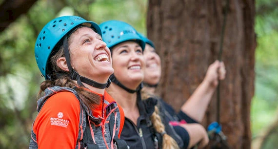 Three people wearing helmets and outdoor gear look upwards, smiling, possibly engaged in an outdoor adventure or climbing activity in a forest area.