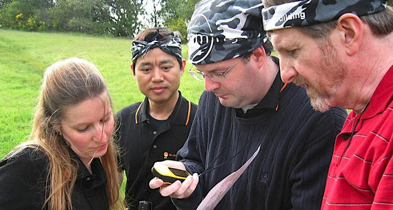 Four people are standing in a grassy area, closely examining a GPS device, likely for navigation or geocaching activities in the outdoors.