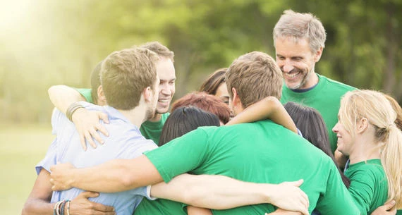 A group of people are standing outdoors, embracing each other in a group hug, smiling and appearing to enjoy their moment together.