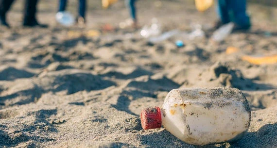 A littered plastic bottle on the sandy beach with blurred people and more trash in the background, indicating environmental pollution.