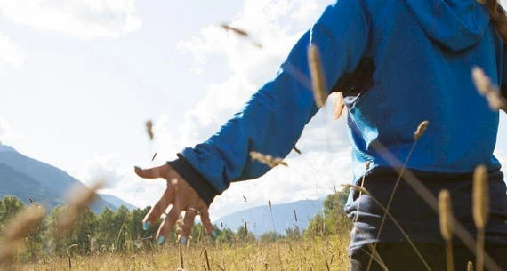 A person in a blue jacket extends an arm while standing in a field with tall grass and mountains in the background under a partly cloudy sky.