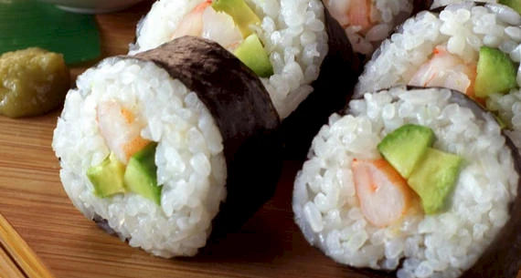 The image shows sushi rolls with rice, seaweed, shrimp, and avocado slices, served on a wooden board with a small amount of wasabi.