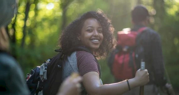 A young woman is smiling while hiking in a forest, carrying a backpack. Others with backpacks and walking sticks are in the background.