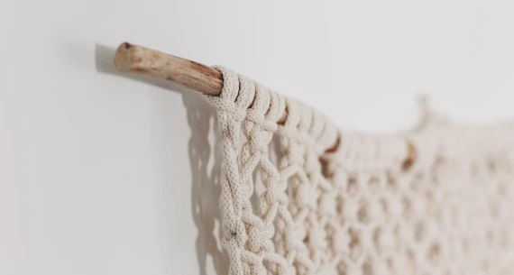 The image shows a close-up of a macramé wall hanging, with intricate knots, attached to a wooden dowel on a white wall.