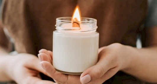 A person is holding a lit white candle in a glass jar, creating a warm and cozy ambiance.