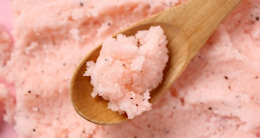 A wooden spoon holds pink exfoliating scrub with small black particles, likely for skincare.