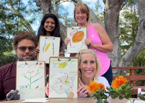 People posing outdoors with finished drawings on clipboards; three women and one man show art pieces, flowers on table, trees in background. Top it at 140 characters, always ending the sentence.