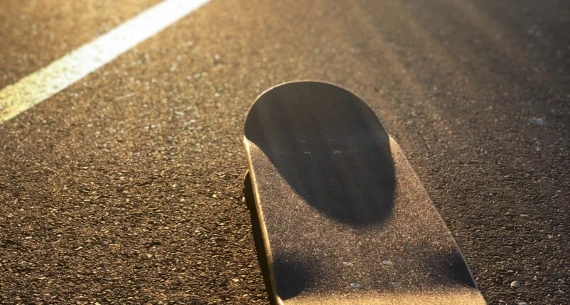 The image shows a skateboard positioned on an asphalt road, with sunlight casting shadows on the board and ground.