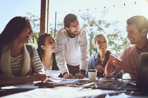 A group of five people are gathered around a table, smiling and engaged in conversation, likely in a collaborative or working environment.